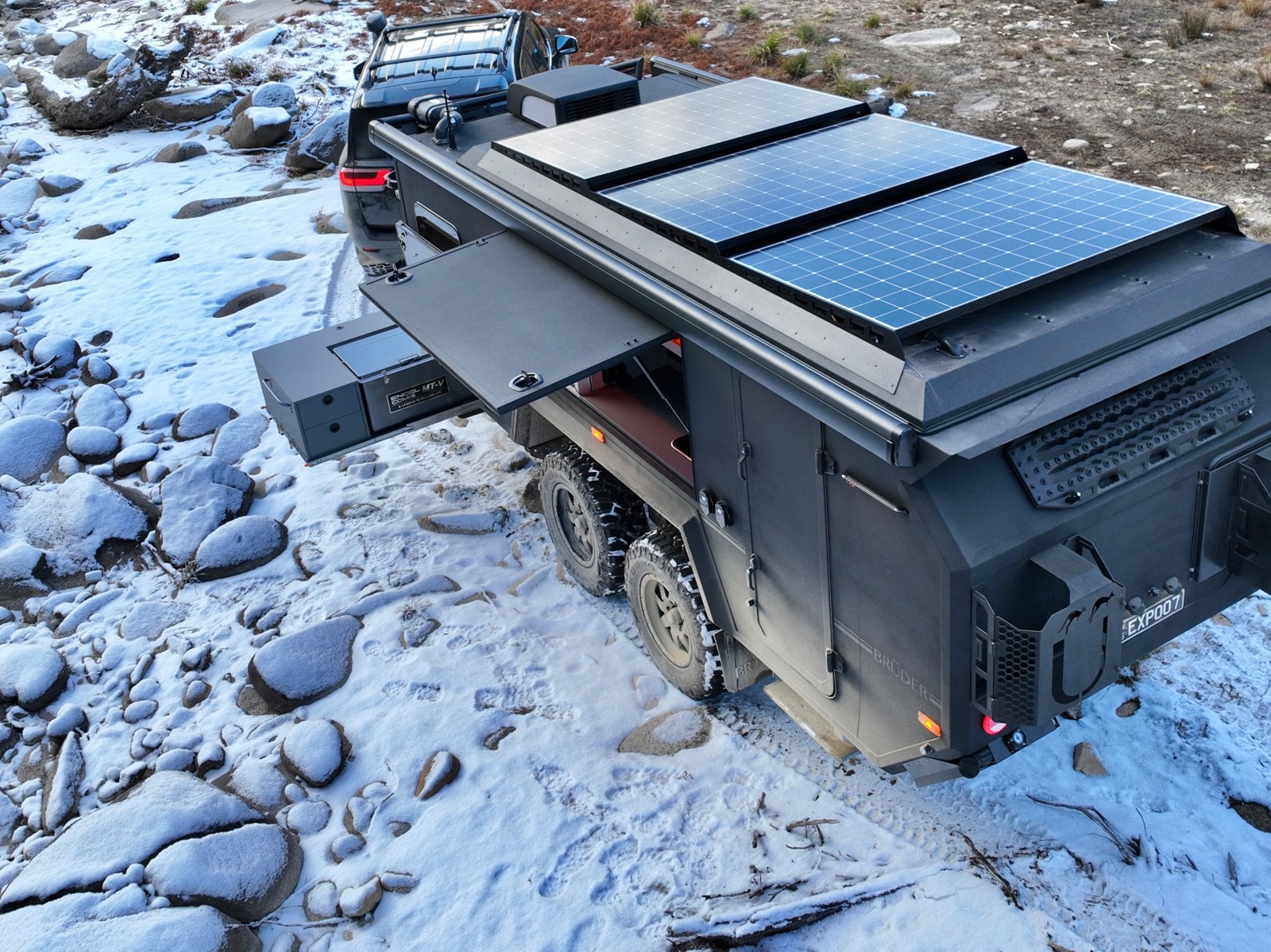 Aerial view of a Bruder expedition trailer with roof solar array on a snow-covered riverbed